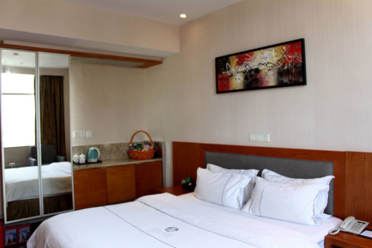 A hotel room with a large bed, a wooden headboard, a bedside table with a phone, and a wall-mounted abstract painting.