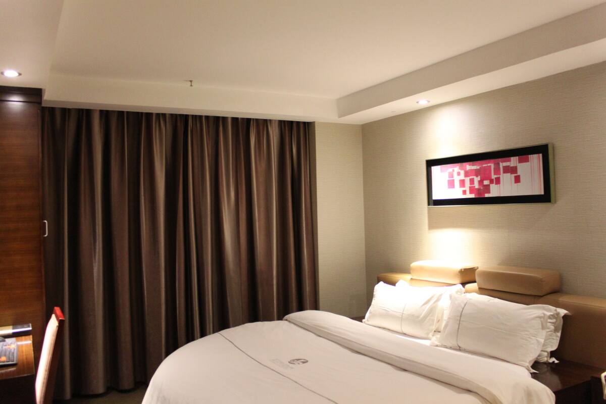 A hotel room with a large bed, a headboard, a nightstand, a lamp, and a wall-mounted artwork.