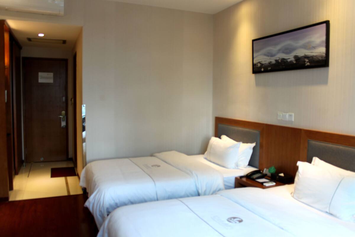 A hotel room with two beds, a wooden headboard, a nightstand with a phone and a tray, and a painting on the wall.
