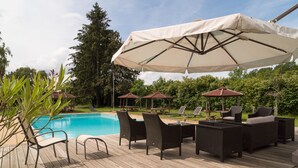 Une terrasse en bois avec des meubles d’extérieur et des parasols, une piscine et de la verdure en arrière-plan.