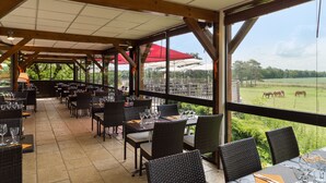 Un restaurant en plein air, doté de tables et de chaises, de grandes fenêtres et offrant une vue sur un champ verdoyant où se trouvent des animaux.