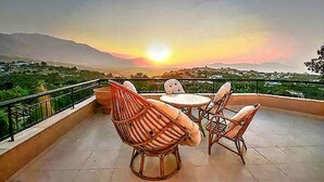 Una terraza con mobiliario de mimbre y vistas a la puesta de sol sobre un paisaje montañoso.