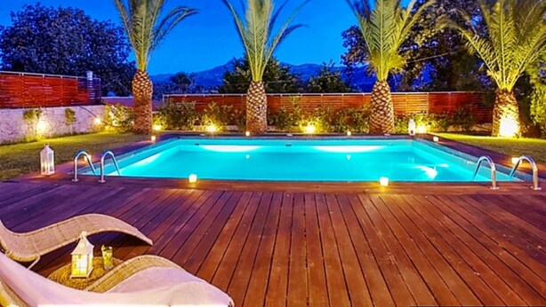 Una piscina con cubierta de madera, palmeras y una montaña al fondo.