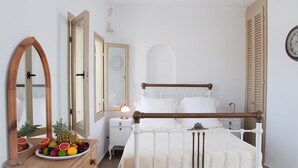 A bedroom with a wooden bed frame, white bedding, a bedside table with a lamp, and a fruit bowl on a dresser. - Lithia Villas (Folegandros)
