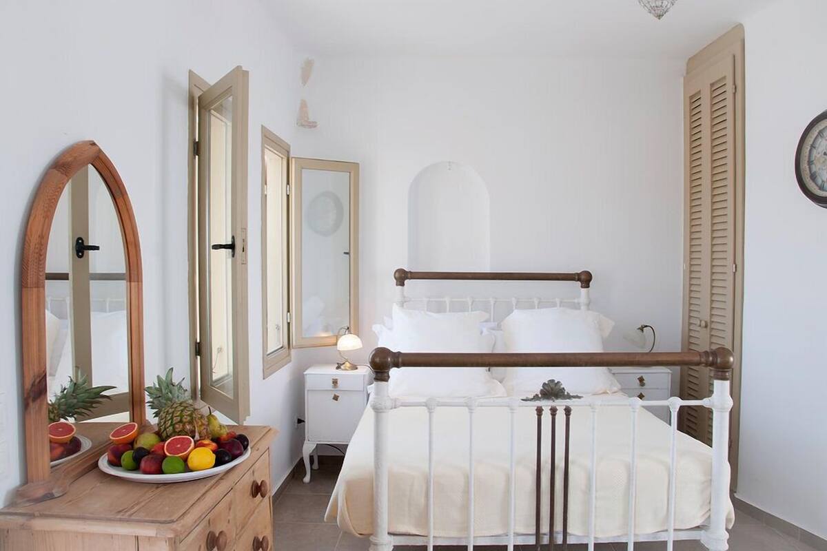 A bedroom with a wooden bed frame, white bedding, a bedside table with a lamp, and a fruit bowl on a dresser.