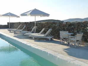 A poolside area with lounge chairs and umbrellas, a stone wall, and a clear view of the water.