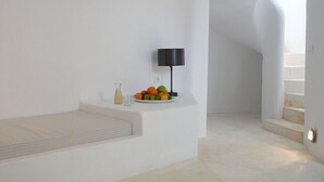 A minimalist room with a white wall, a small table with a fruit bowl, a lamp, and a bench. - Lithia Villas (Folegandros)