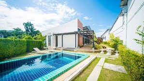 A modern outdoor swimming pool area with a clear blue pool, surrounded by well-maintained greenery and a white building with a covered patio. - ChillHub Hostel (Choeng Thale)