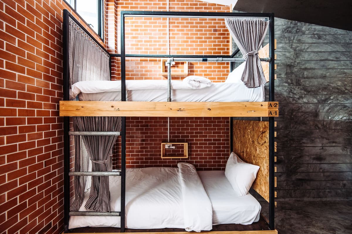 A bunk bed with a wooden frame and metal railings, set against a brick wall with a wooden panel on the right.