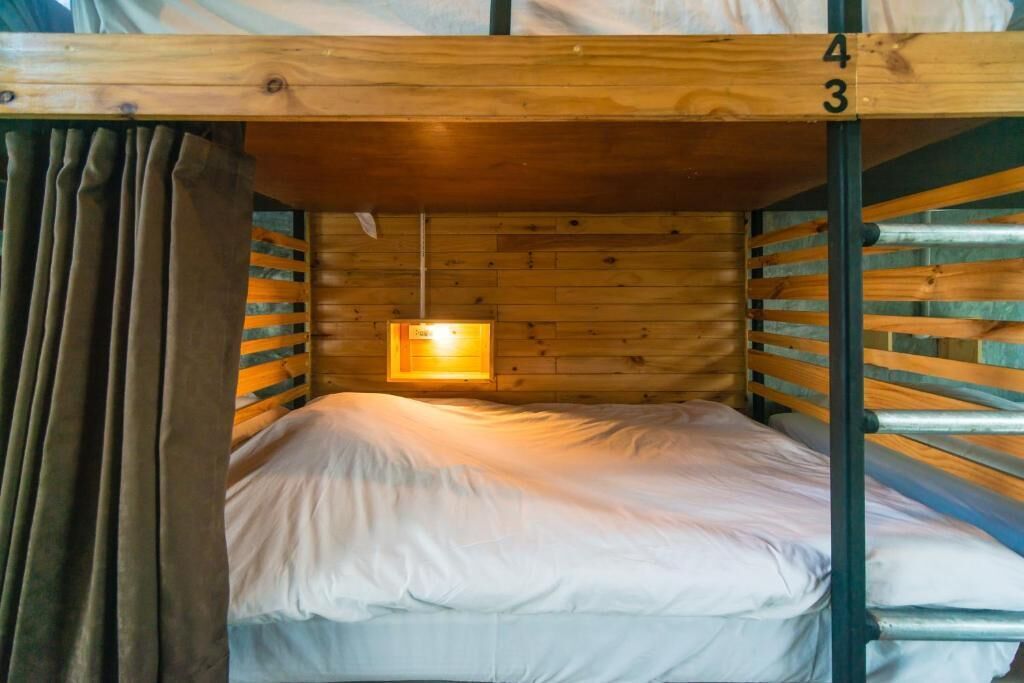 A bunk bed room with a wooden slat wall, a single bed with white bedding, and a small window with a light fixture.