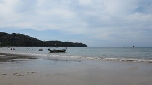 A beach with boats, a distant hill with buildings, and a clear sky. - ChillHub Hostel (Choeng Thale)