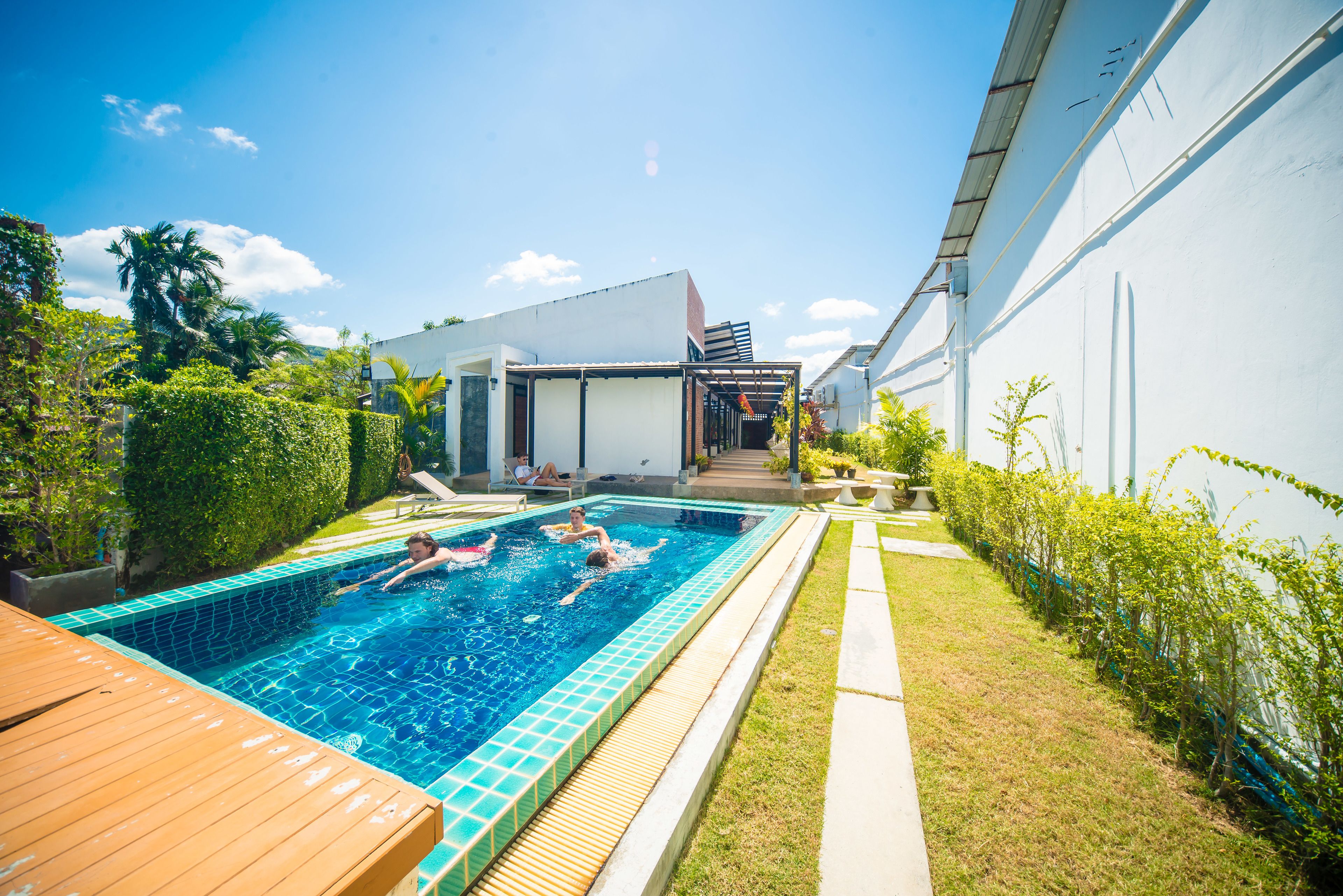 A swimming pool with a wooden deck, surrounded by greenery and a white building.