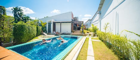 A swimming pool with a wooden deck, surrounded by greenery and a white building.