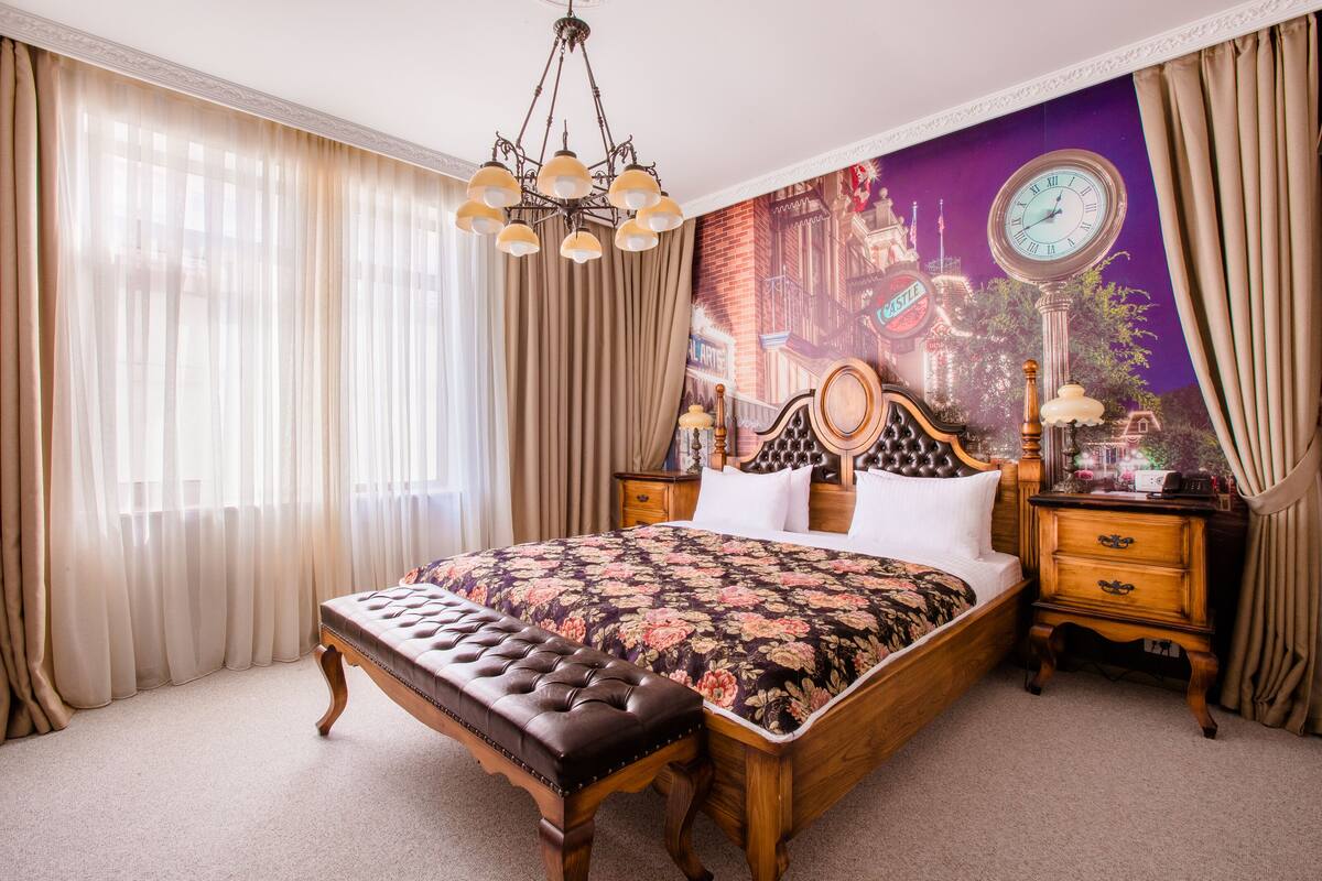 Une chambre spacieuse avec un grand lit, un banc en bois, une horloge classique et une fresque représentant un paysage urbain.