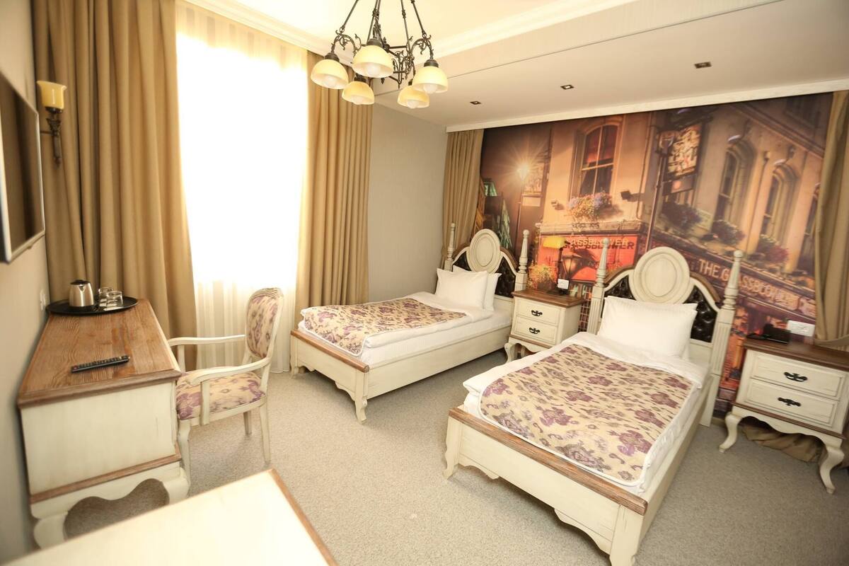 Une chambre d’hôtel avec deux lits, un bureau, une chaise, une commode et une grande fenêtre avec des rideaux.