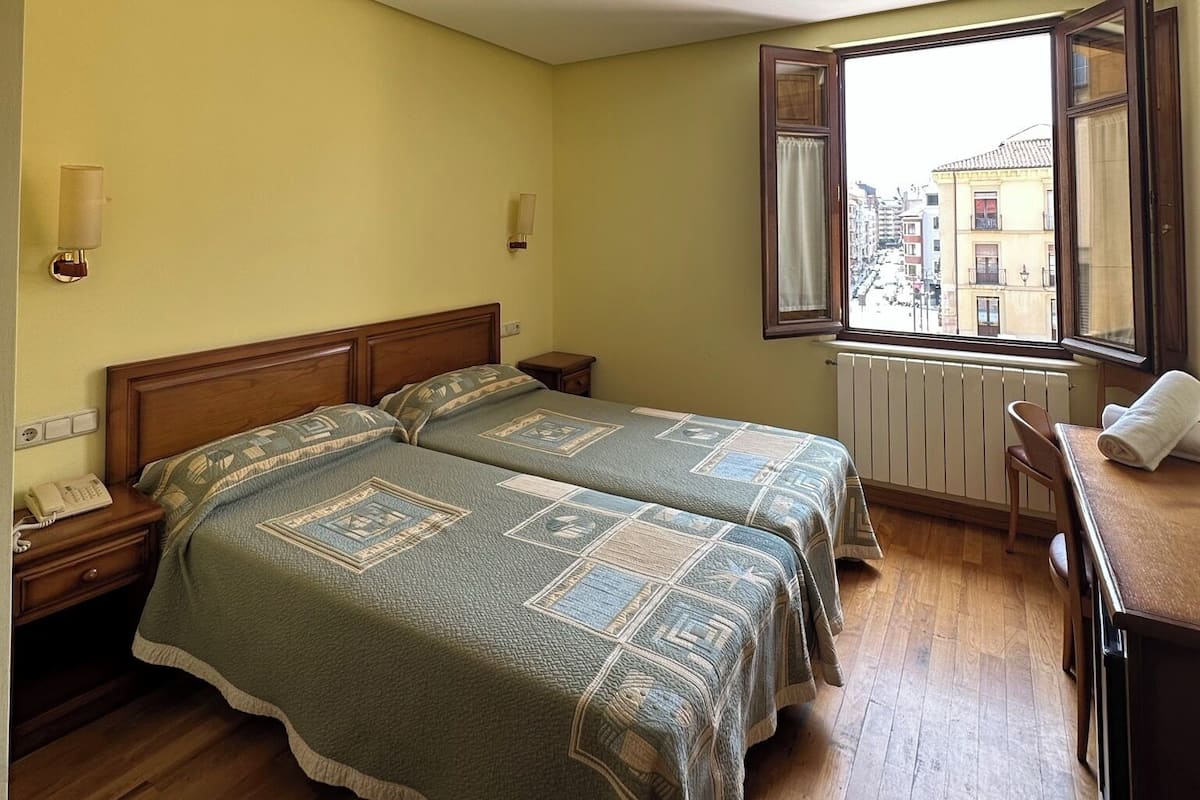 A hotel room with two beds, wooden flooring, a window with a view of buildings, and a desk with a chair.