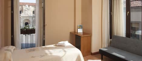 A hotel room with a bed, a sofa, a small wooden table, and a view of a building through a balcony door.