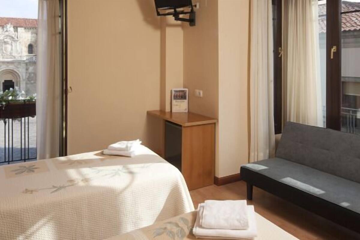 A hotel room with a bed, a sofa, a small table, and a view of a building through a glass door.