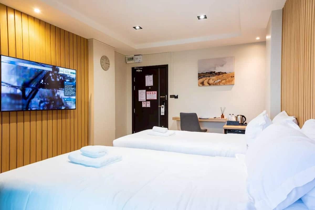 A hotel room with two beds, a desk, a chair, a flat-screen TV mounted on the wall, and a painting on the wall.
