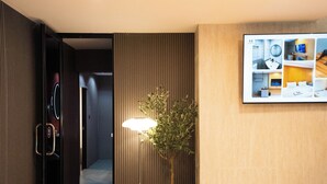 Ett hotellrum med en säng, en tv som är monterad på väggen, en krukväxt och en korridor som leder till ett annat rum.