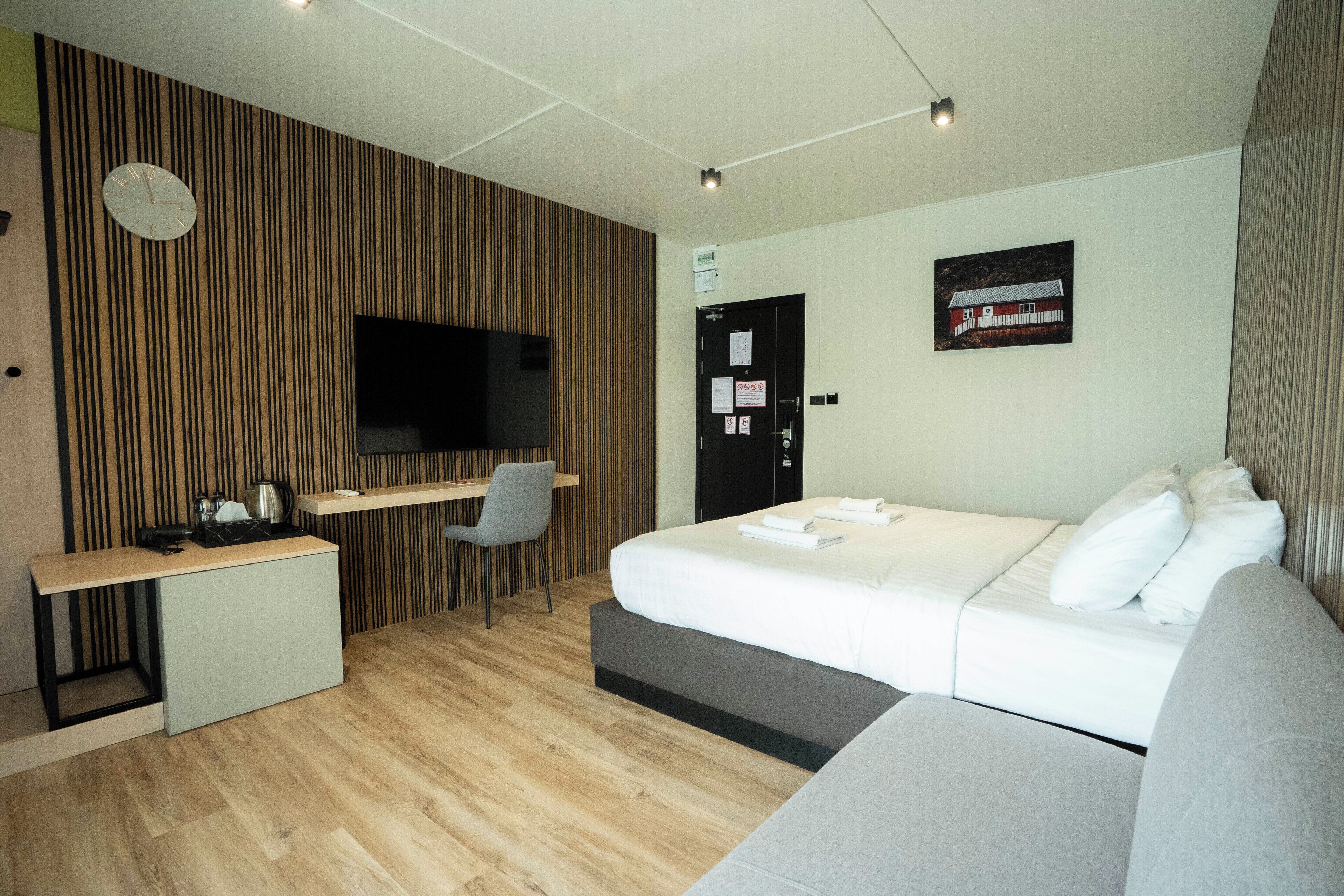 A modern hotel room with a wooden panel wall, a flat-screen TV, a bed with white bedding, a grey sofa, and a desk with a chair.
