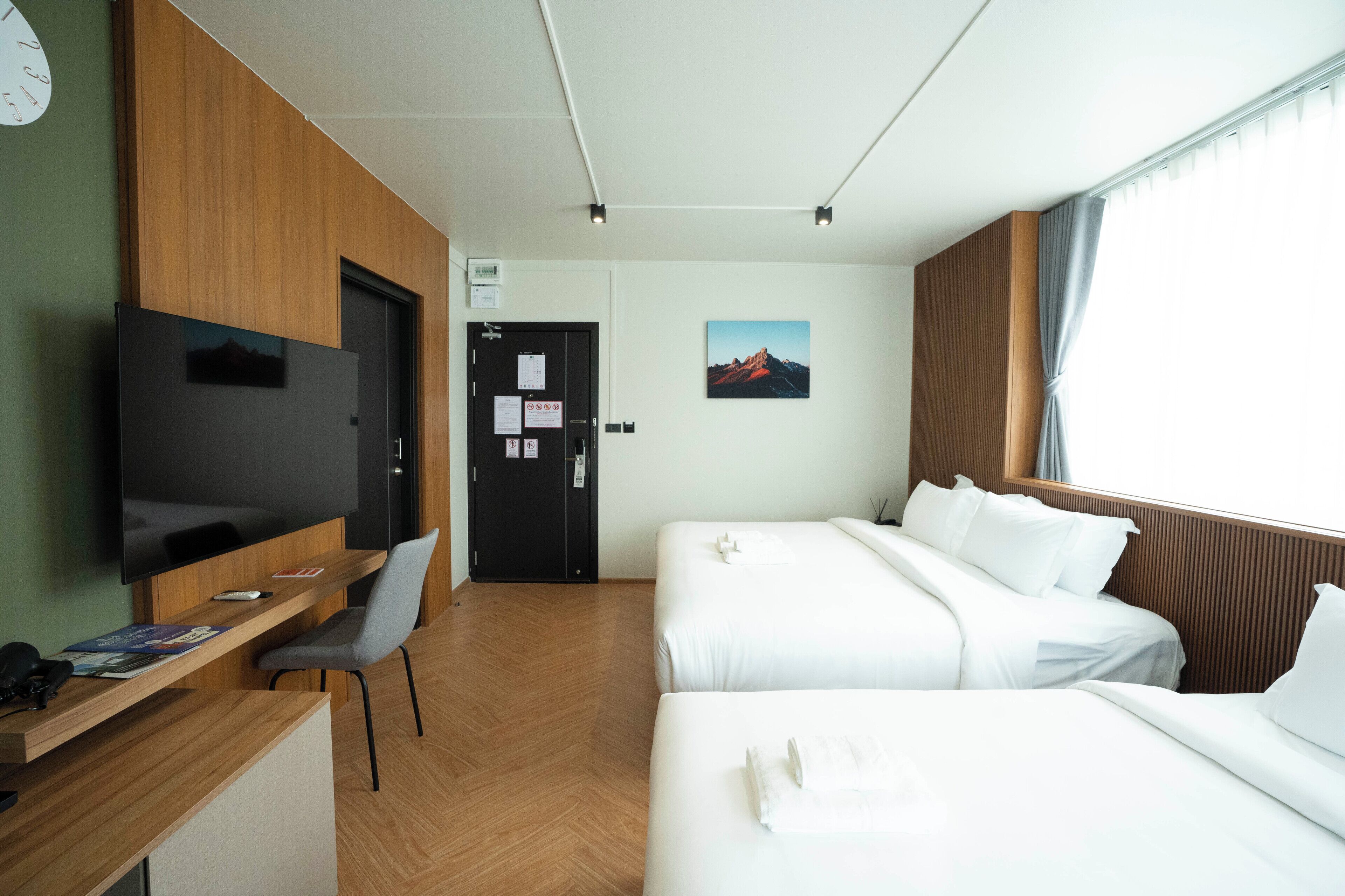 A hotel room with a wooden desk, a flat-screen TV, two beds, a painting on the wall, and a window with curtains.