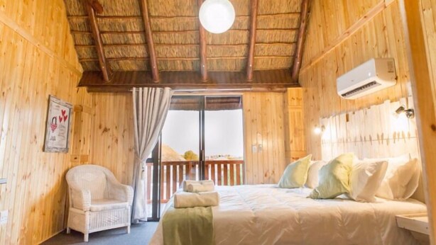 A wooden cabin bedroom with a bed, a chair, and a window with curtains. - Kalahari Lion's Rest (Upington)