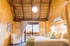 A wooden cabin bedroom with a bed, a chair, and a window with curtains. - Kalahari Lion's Rest (Upington)