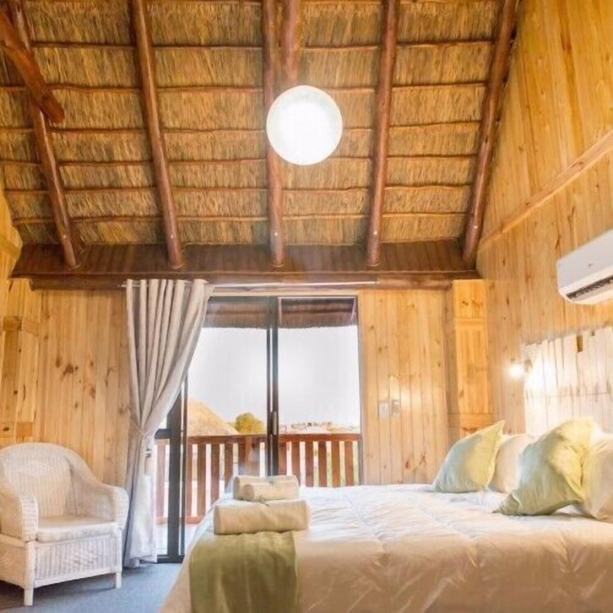 A wooden cabin room with a bed, a chair, and a window with curtains.