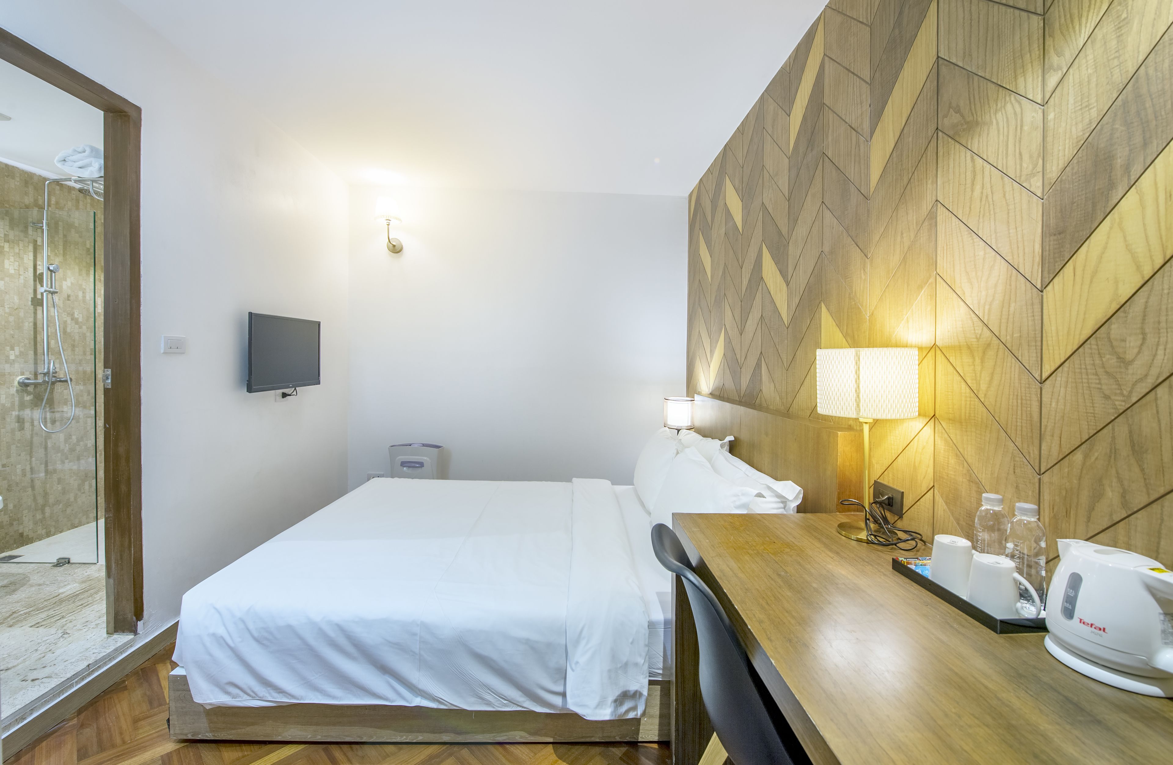 A hotel room with a bed, a desk with a lamp, a TV, and a shower area.