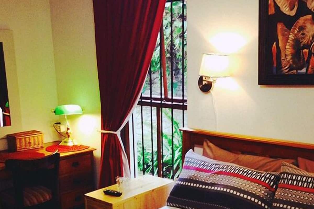 A hotel room with a bed, a nightstand, a lamp, a window with curtains, and a wall-mounted light.
