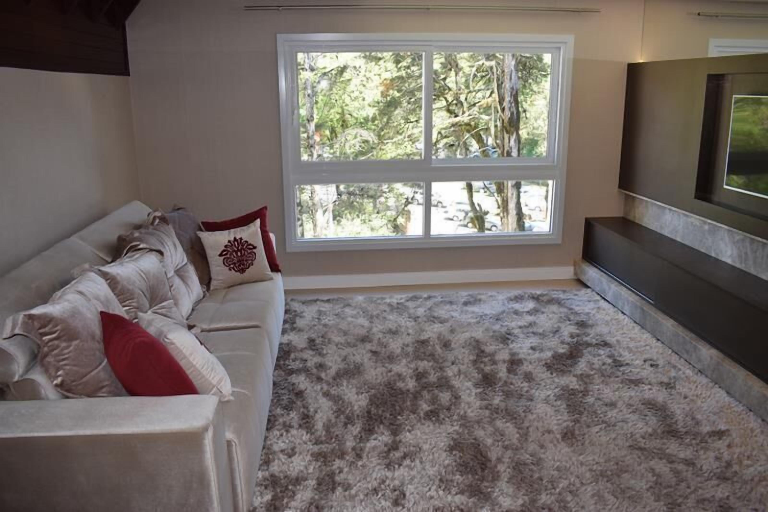 A living room with a white sofa, a large window with a view of trees, and a flat-screen TV mounted on the wall.