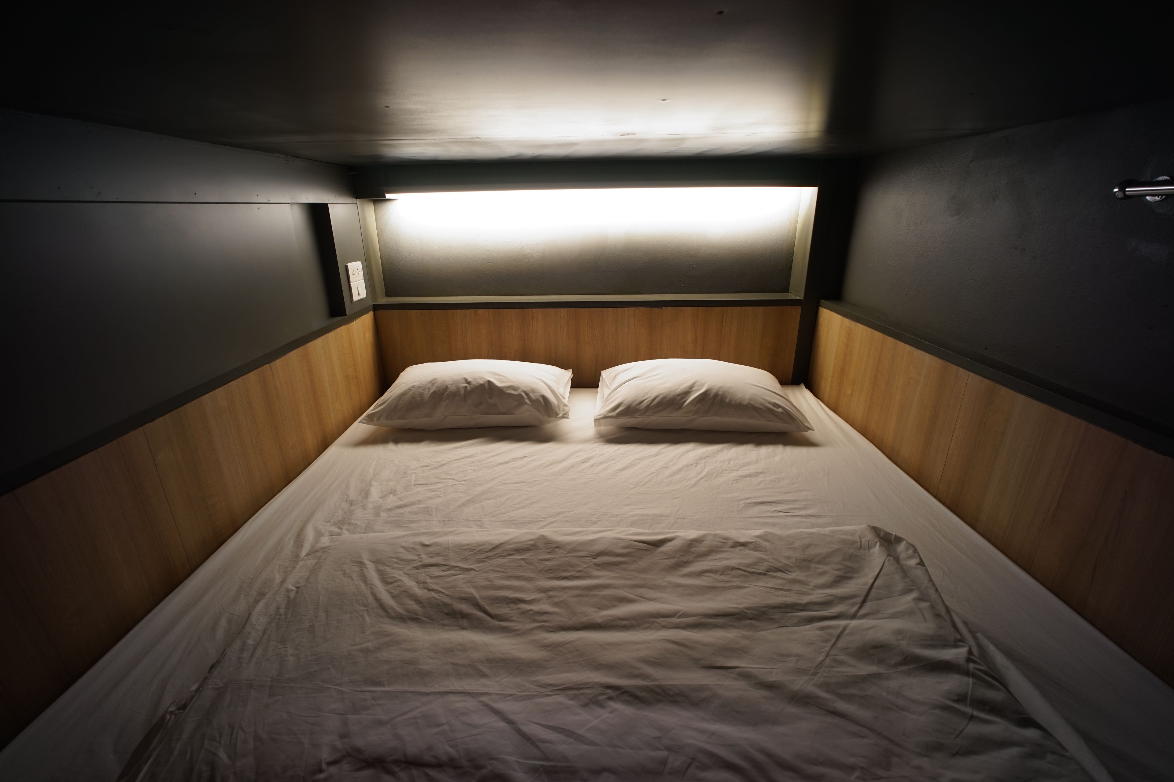 A narrow, dimly lit room with a single bed, wooden headboard, and a small window providing minimal natural light.