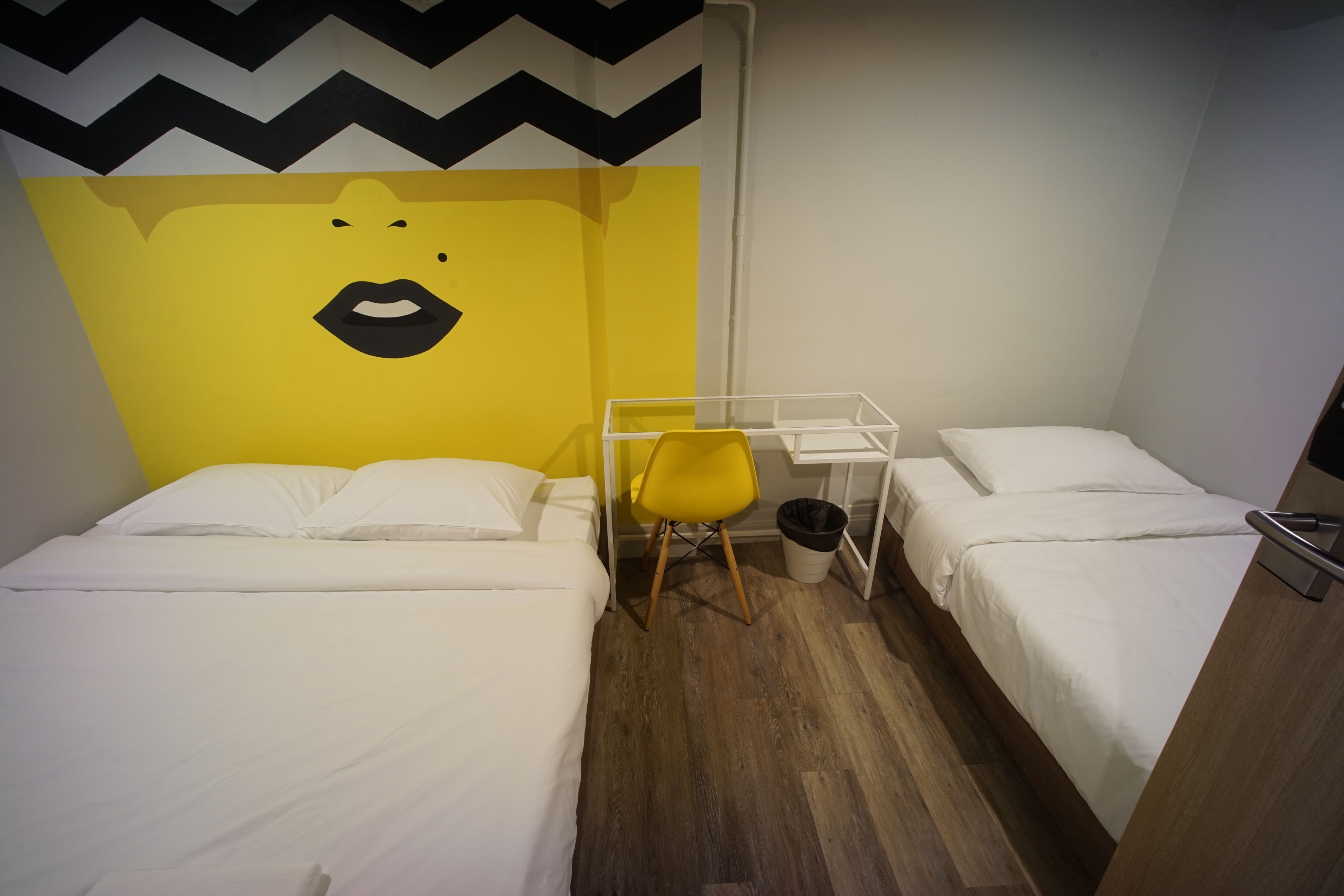 Une chambre d’hôtel avec deux lits, une chaise jaune, un bureau blanc et un mur orné d’un motif noir et blanc.