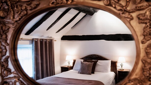 Un dormitorio con una cama grande, dos mesitas de noche con lámparas, un ventanal con cortinas y un espejo con marco decorativo.