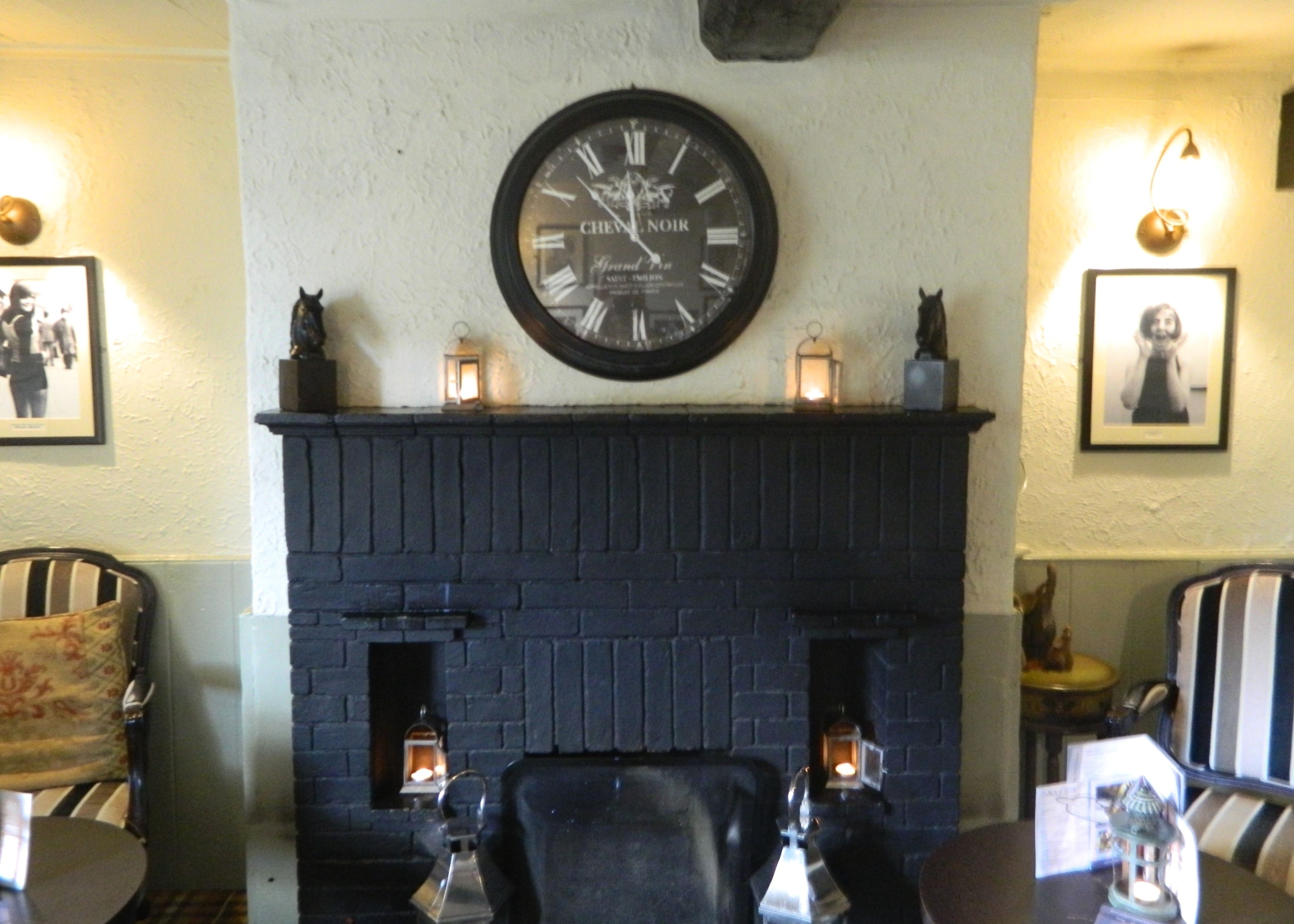 Una habitación con chimenea de ladrillo negro, un reloj de pared grande y dos faroles encendidos.