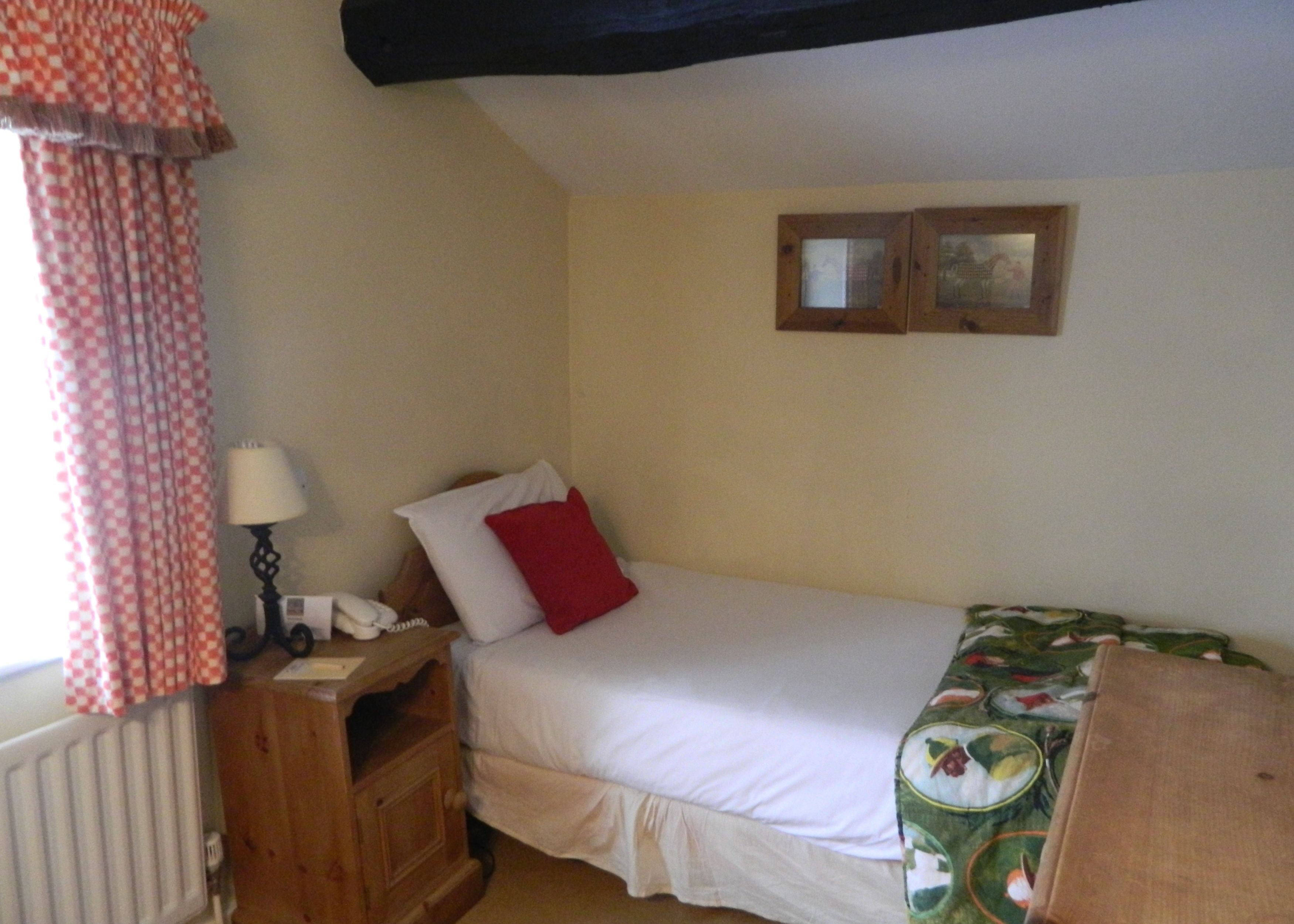 Uma cama de solteiro com um edredom branco e um travesseiro vermelho, uma mesa de cabeceira de madeira com uma luminária, um radiador e uma janela com cortinas xadrez.