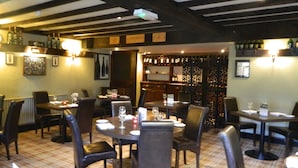 Interior de restaurante con vigas de madera, suelo a cuadros, mesas con sillas, botellas de vino en estanterías y cuadros en las paredes.
