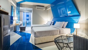 Una camera d'albergo moderna con una parete blu a contrasto, un letto con biancheria bianca, un tavolino con una lampada e un angolo cottura.