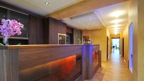 Una moderna recepción con encimera de madera e iluminación tenue, un florero con flores moradas, y un pasillo con suelo de madera que conduce a una habitación luminosa.