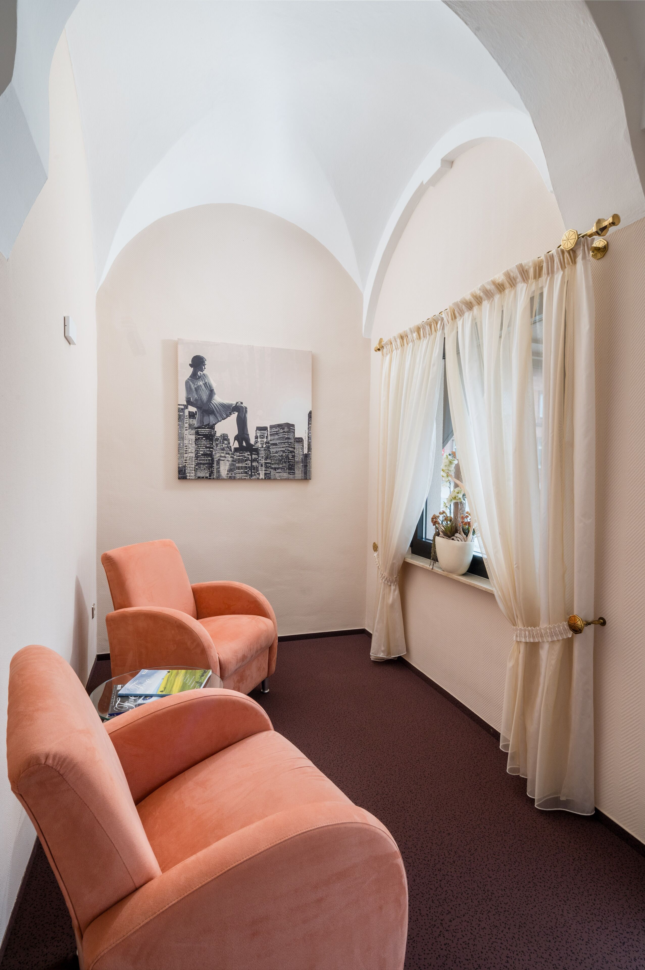 Ein Zimmer mit einem pfirsichfarbenen Sessel, einem Stadtbildgemälde und einem Fenster mit transparenten Vorhängen.