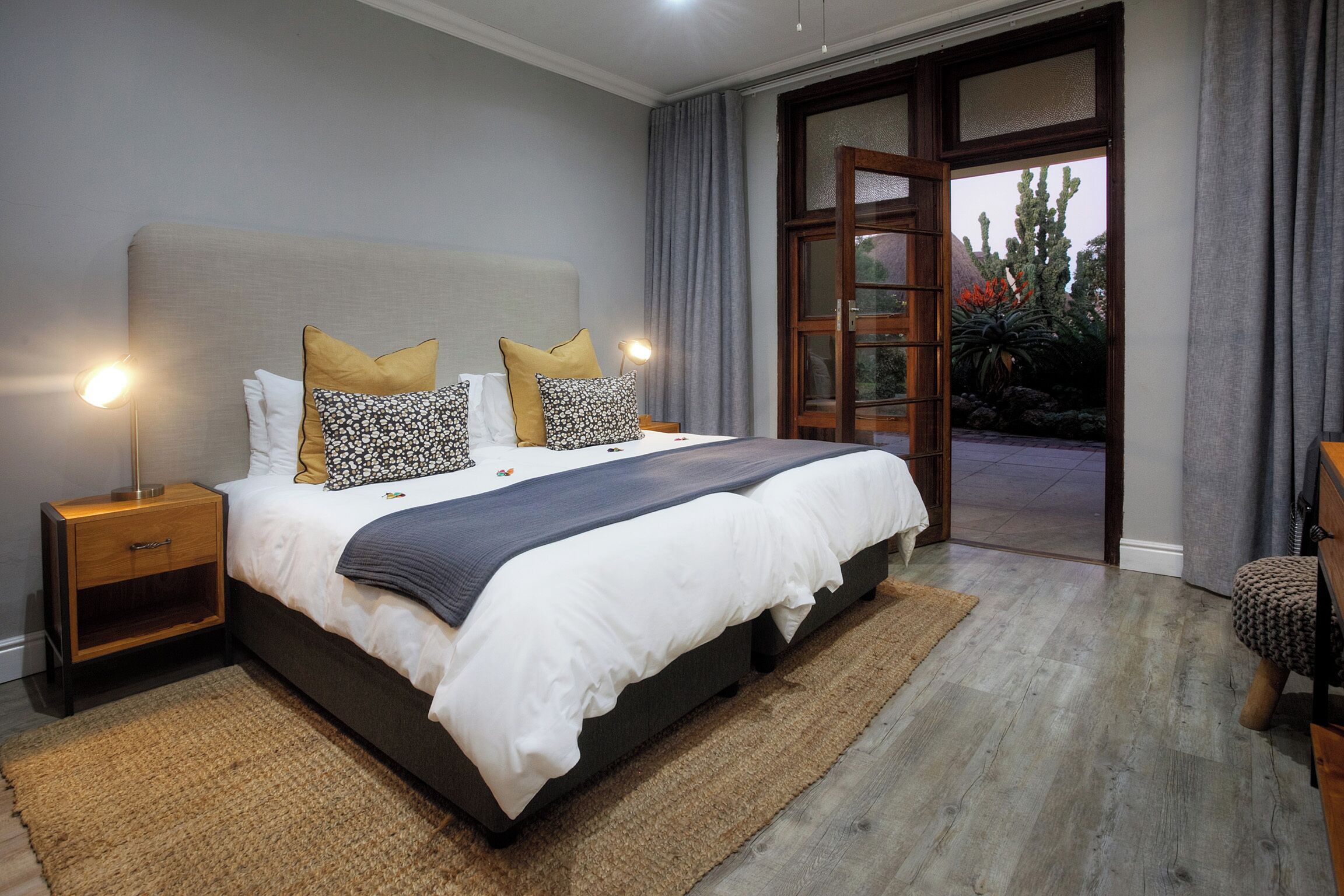 A bedroom with a large bed, two bedside tables, a nightstand, and a view of a garden through a glass door.