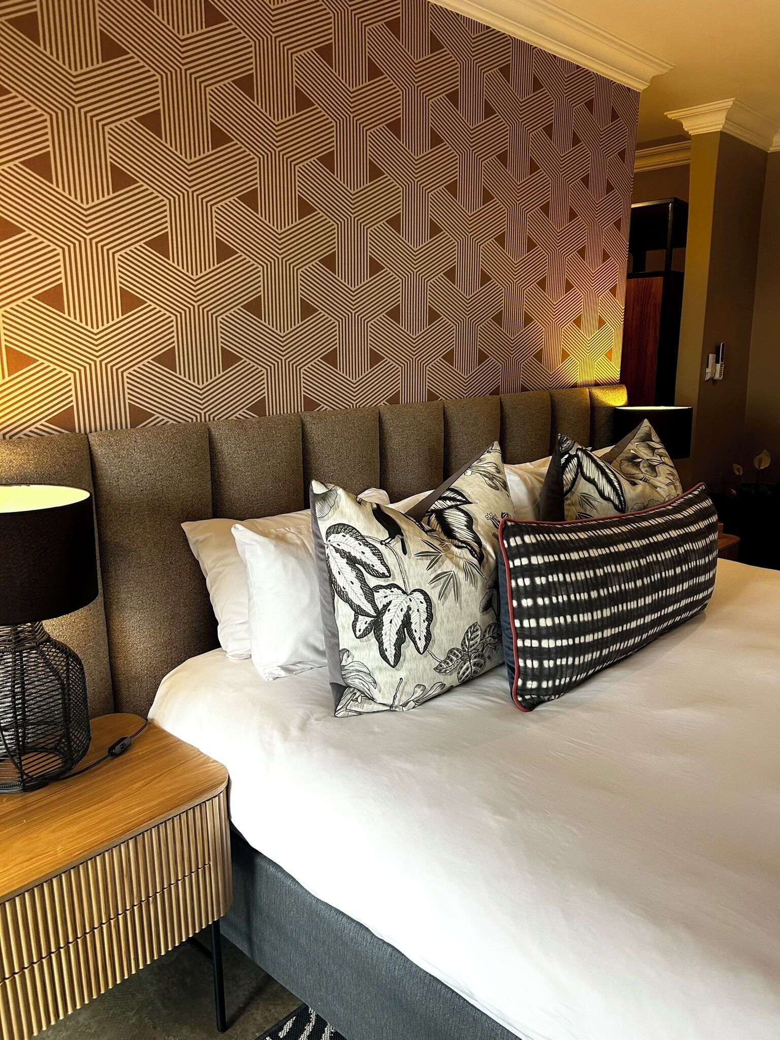 A hotel room with a patterned wallpaper, a bed with multiple pillows, a bedside table with a lamp, and a nightstand.