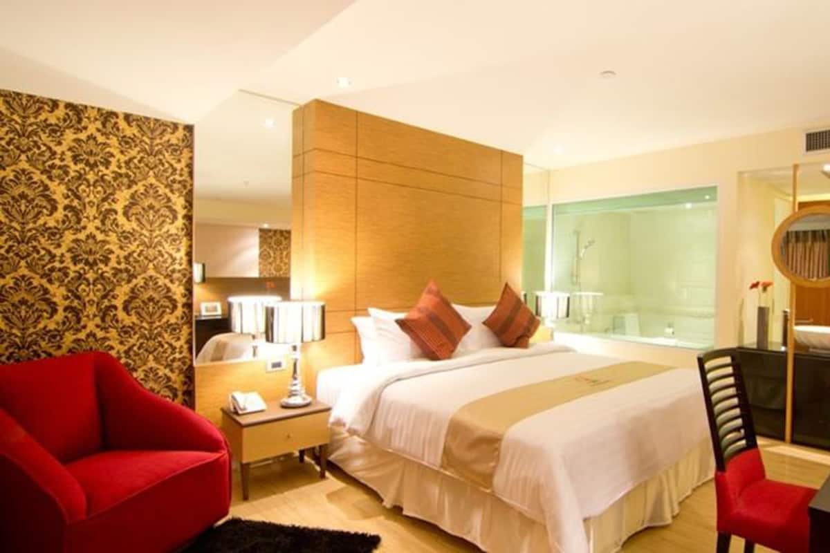 A hotel room with a large bed, a red armchair, a desk with a lamp, and a large mirror.