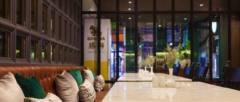 A modern hotel lobby with a leather sofa, a long dining table, and a view of the city through large windows.