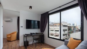 A modern living room with a grey sofa, a small desk, and a chair. A flat-screen TV is mounted on the wall, and there is a large window with curtains. - Oriental Heritage Hotel (Bangkok)