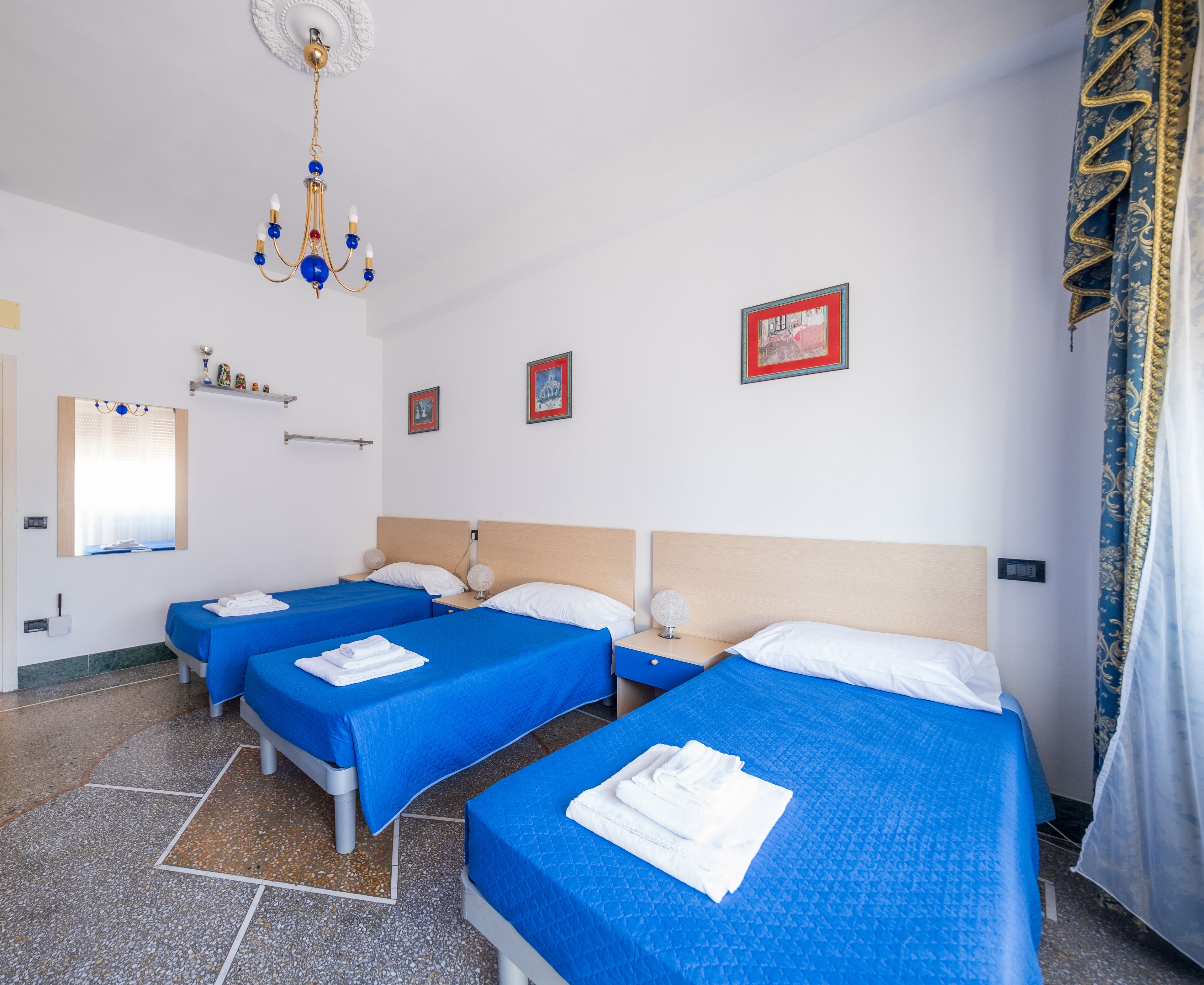 A hotel room with three single beds, each with a blue bedspread and white pillows. There is a chandelier, a mirror, and framed pictures on the wall.