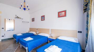 A hotel room with three single beds, each with a blue bedspread and white pillows. There is a chandelier, a mirror, and framed pictures on the wall.