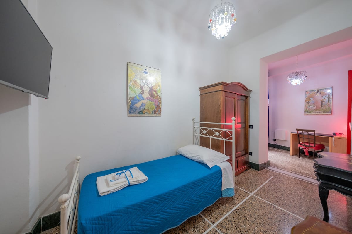 A bedroom with a single bed, a wooden wardrobe, a desk with a chair, and two paintings on the walls.