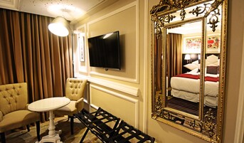 A hotel room with a television, a large ornate mirror, a tufted armchair, a small round table, and a bed with a red blanket.